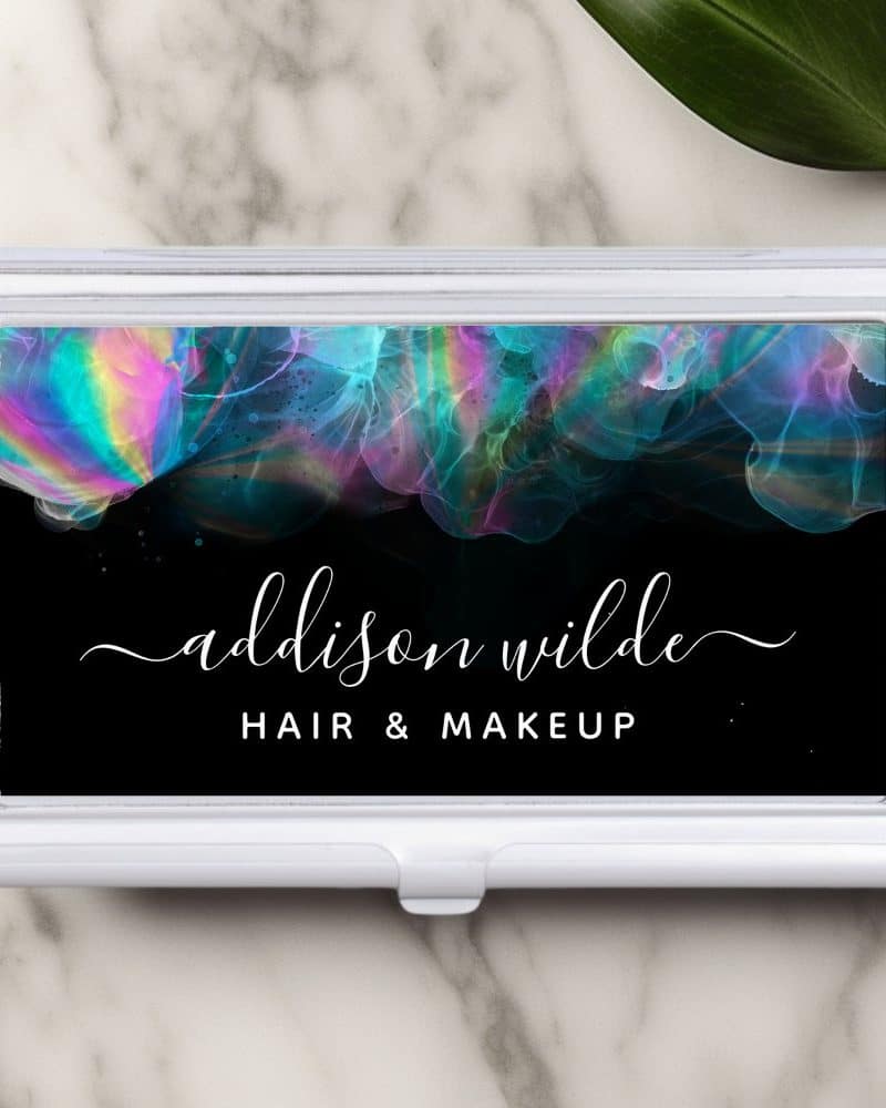 A white business card holder displays a black background with iridescent teal, pink, and purple abstract designs. The name Addison Wilde, Hair & Makeup is written in white script.