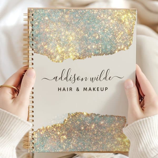 Two Hands Hold A Spiral-Bound Notebook With A Sparkly Gold And Teal Cover. The Name Addison Wilde Hair &Amp; Makeup Is Printed On The Cover.