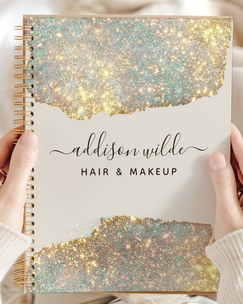 Two hands hold a spiral-bound notebook with a sparkly gold and teal cover. The name Addison Wilde Hair & Makeup is printed on the cover.