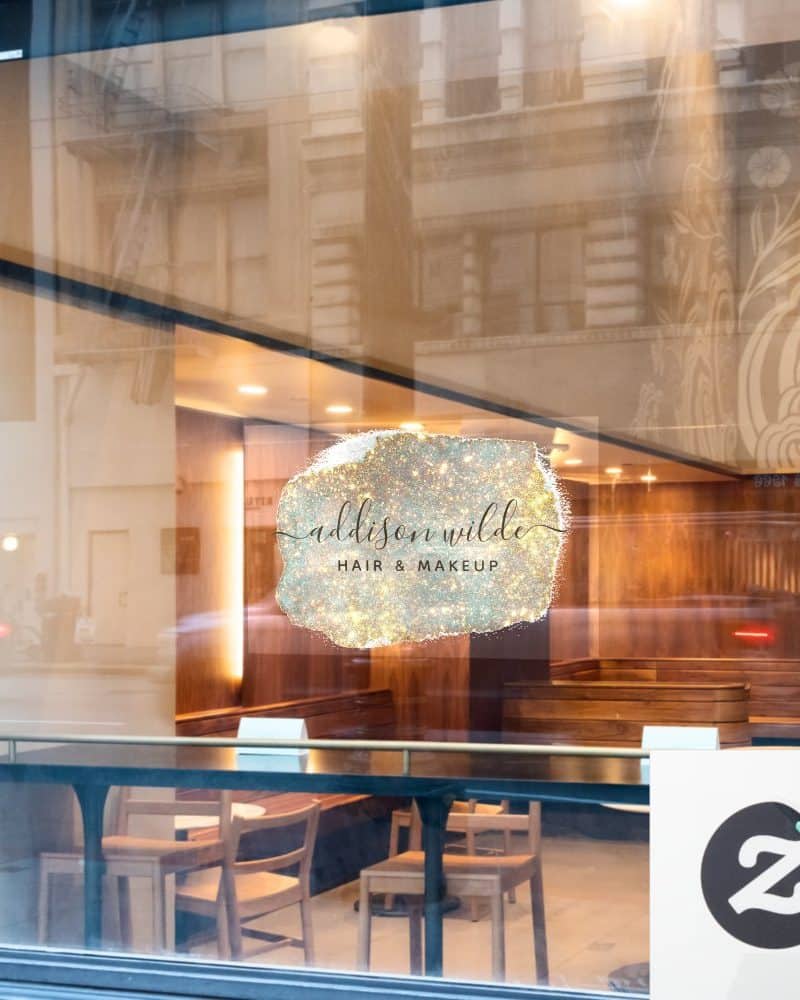 A salon logo on a storefront window. The logo is a sparkly, light-colored, abstract shape with the words Addison Wilde Hair & Makeup in elegant script.