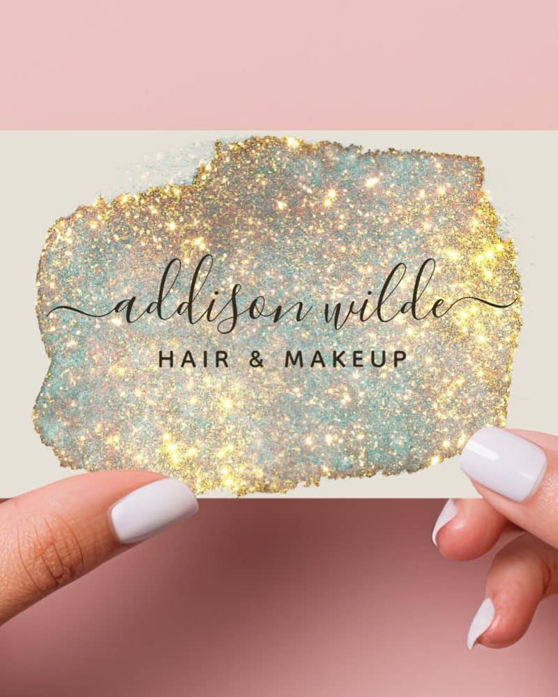 Two hands hold up a business card featuring a sparkly gold and teal design. The name Addison Wilde Hair & Makeup is displayed on the card.
