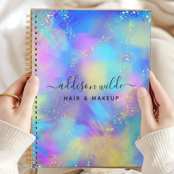 Two Hands Hold A Spiral-Bound Notebook With A Pastel Holographic Cover. The Name Addison Wilde Hair &Amp; Makeup Is Printed On The Cover.