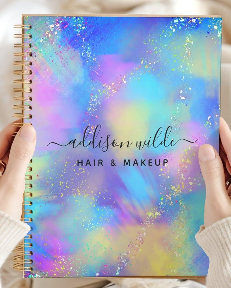 Two hands hold a spiral-bound notebook with a pastel holographic cover. The name Addison Wilde Hair & Makeup is printed on the cover.