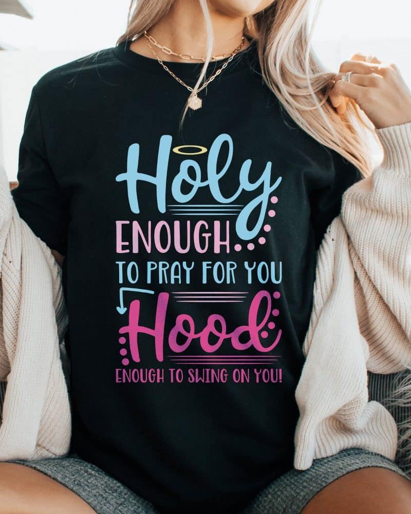 Woman wearing a black t-shirt that says Holy Enough to Pray for You, Hood Enough to Swing on You.