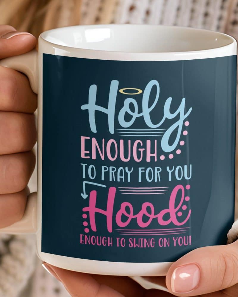 A white coffee mug with a dark blue design that says Holy Enough to pray for you, Hood Enough to swing on you.