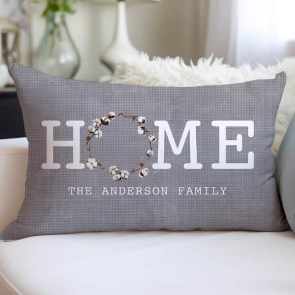 Gray Rectangular Pillow With The Word Home In White Sans-Serif Font, A Cotton Wreath In The Center Of The Word, And The Anderson Family Written Below.