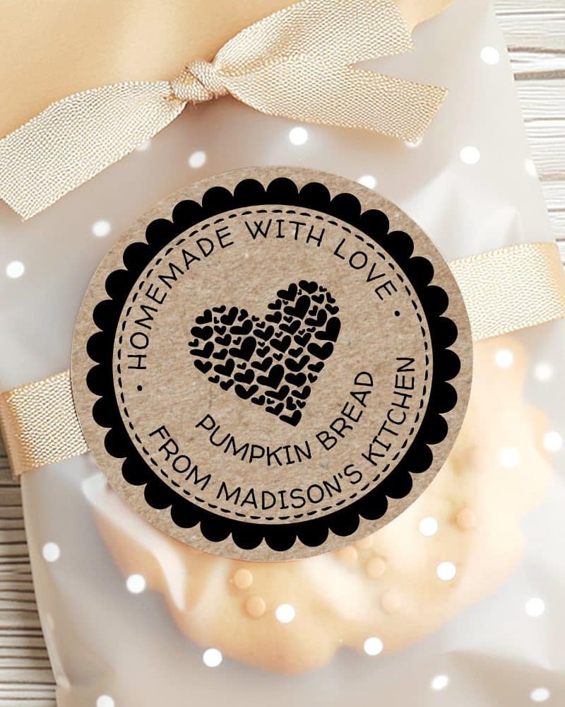 A round label with a black border and a kraft paper background shows a heart made of smaller hearts. The text reads Homemade with Love, Pumpkin Bread, from Madisons Kitchen. The label is attached to a cellophane bag of baked goods.