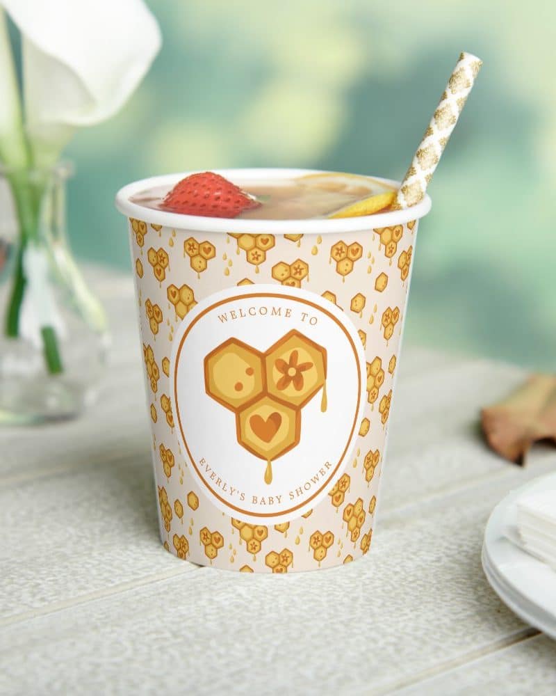A paper cup with a honey-comb themed baby shower design, filled with a beverage and a straw.