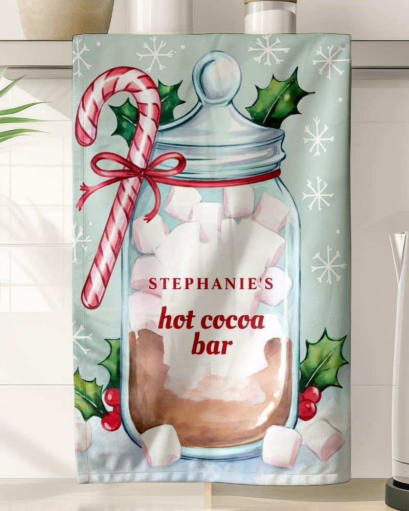 A kitchen tea towel showing a mason jar of hot cocoa with marshmallows, a candy cane, and holly. The text Stephanie's Hot Cocoa Bar is also on the towel.