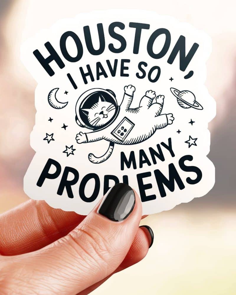 A hand holds a sticker featuring a cat in an astronaut suit floating in space, with the text Houston, I have so many problems.