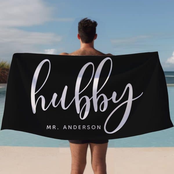 A Man Holds A Black Beach Towel Behind His Back. The Towel Says Hubby In Elegant Script Lettering With Mr. Anderson Below It.