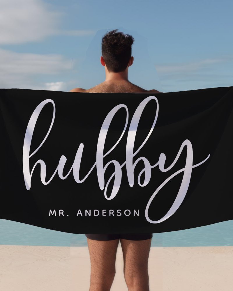 A man holds a black beach towel behind his back. The towel says Hubby in elegant script lettering with Mr. Anderson below it.