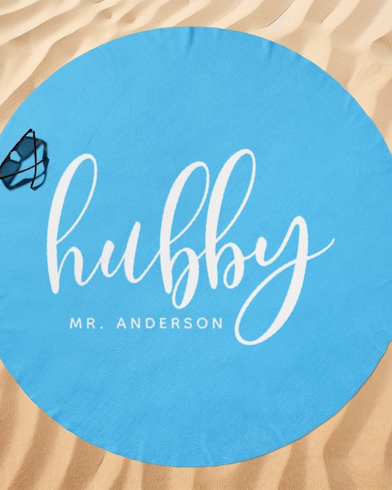 Light blue round beach towel with the word hubby in white script lettering and Mr. Anderson below it. A pair of sunglasses rests on the towel.
