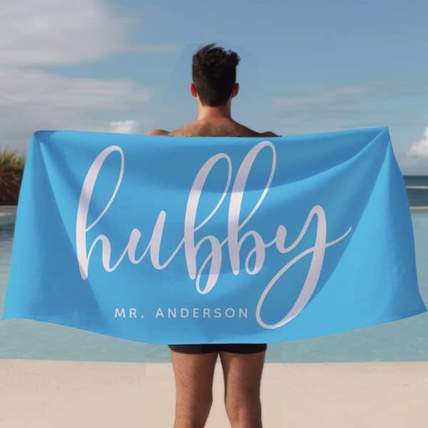 A Man Holds A Bright Blue Beach Towel Behind His Back. The Towel Says Hubby In A Stylized White Script Font, With Mr. Anderson Below It.