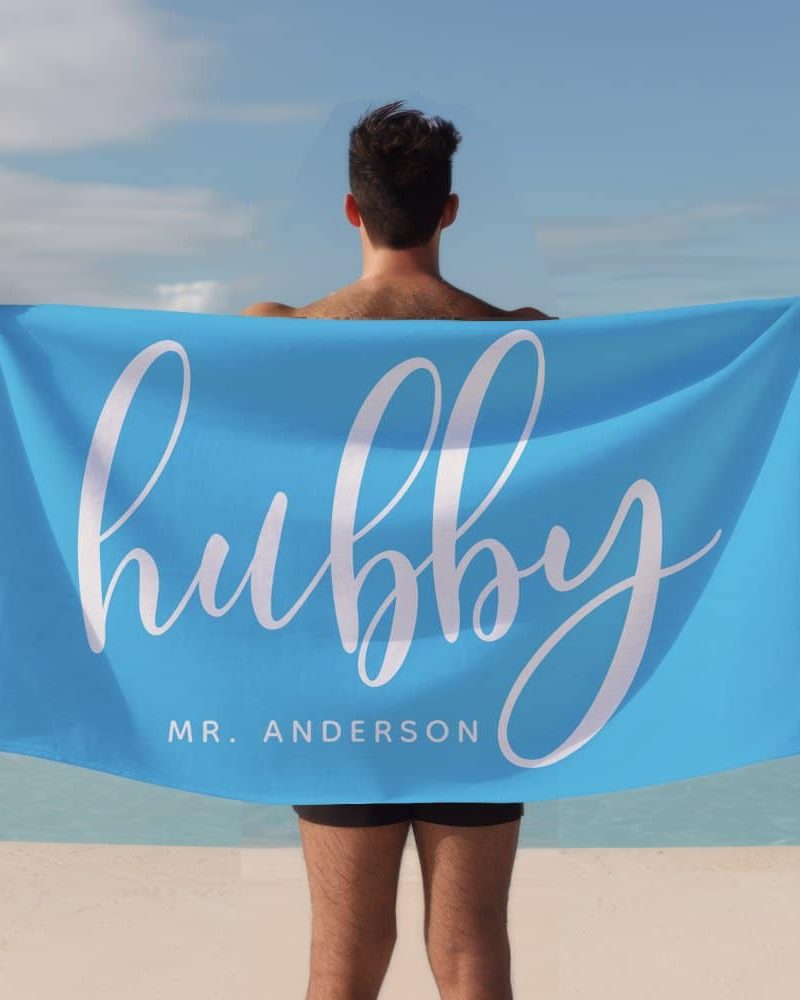 A man holds a bright blue beach towel behind his back. The towel says hubby in a stylized white script font, with Mr. Anderson below it.