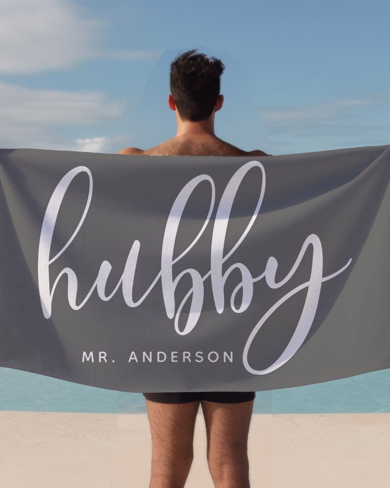 A man holds a gray beach towel behind him. The towel says Hubby in stylized white lettering, with Mr. Anderson below.