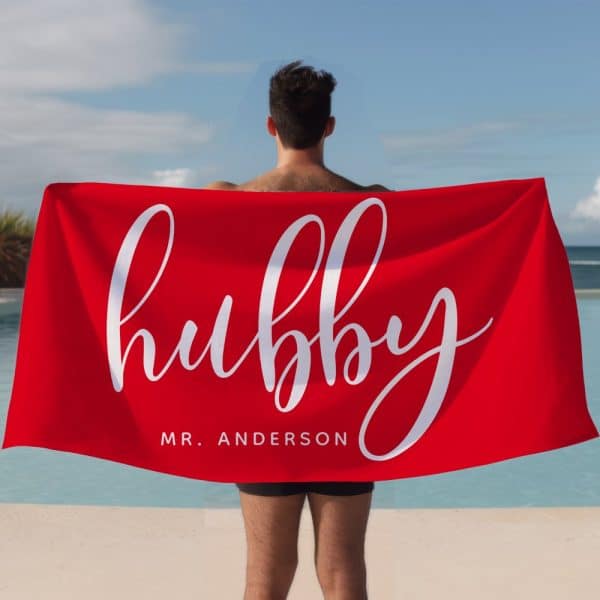 A Man Holds A Large Red Beach Towel Behind His Back. The Towel Says Hubby In A Stylized White Script Font And Mr. Anderson In A Smaller Font Below.