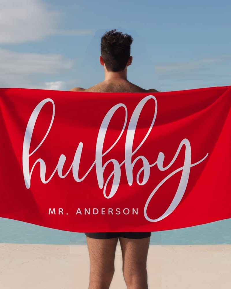 A man holds a large red beach towel behind his back. The towel says Hubby in a stylized white script font and Mr. Anderson in a smaller font below.