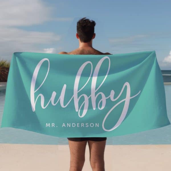 A Man Holds A Teal Beach Towel Behind His Back. The Towel Says Hubby In Stylized White Lettering, With Mr. Anderson Below.