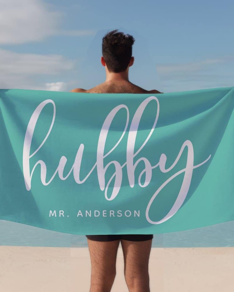A man holds a teal beach towel behind his back. The towel says hubby in stylized white lettering, with Mr. Anderson below.