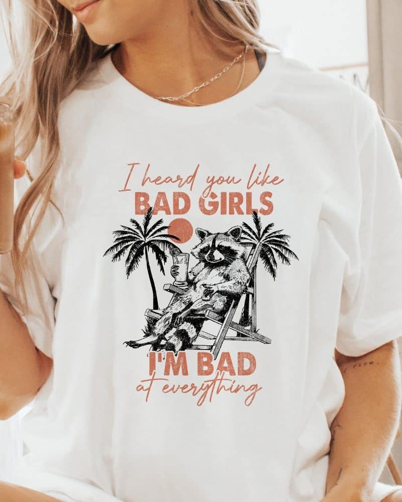 White t-shirt featuring a graphic design of a raccoon lounging in a beach chair with a drink, palm trees, and text that reads, I heard you like bad girls, I'm bad at everything.