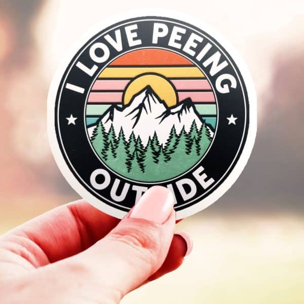A Hand Holds A Round Sticker With A Mountain Scene And The Text I Love Peeing Outside.