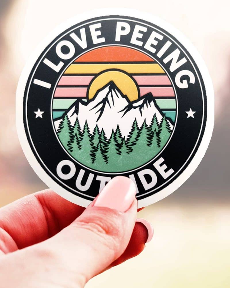 A hand holds a round sticker with a mountain scene and the text I Love Peeing Outside.
