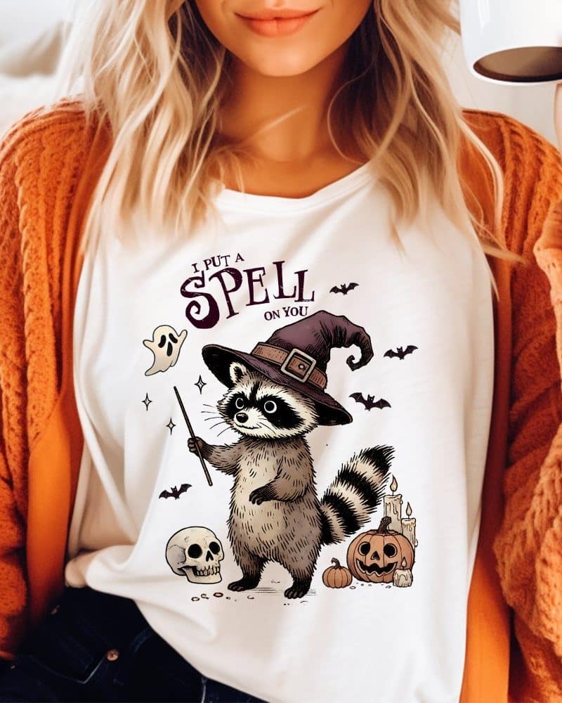 White shirt featuring a cartoon raccoon wearing a witch's hat and holding a magic wand. Halloween themed design includes pumpkins and a skull.
