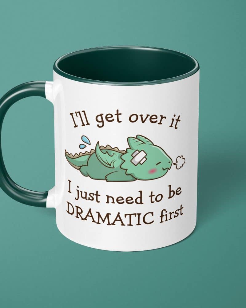 White coffee mug with dark green handle and interior, featuring a cartoon illustration of a small, injured dragon and text that reads, Ill get over it I just need to be DRAMATIC first.