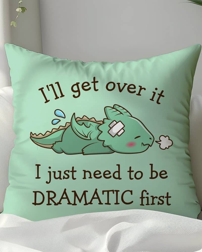 Light green pillow featuring a cartoon illustration of a small, injured dragon lying down. Text on the pillow reads, Ill get over it I just need to be DRAMATIC first.