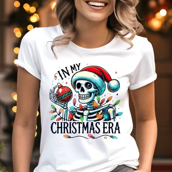 White T-Shirt Featuring A Skeleton Wearing A Santa Hat, Holding An Ornament, Surrounded By Christmas Lights; Text Reads In My Christmas Era.