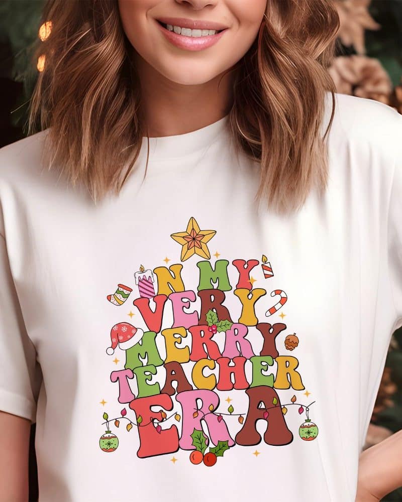 Woman wearing a white t-shirt with a Christmas tree design. The tree is made of colorful letters spelling In My Very Merry Teacher Era.