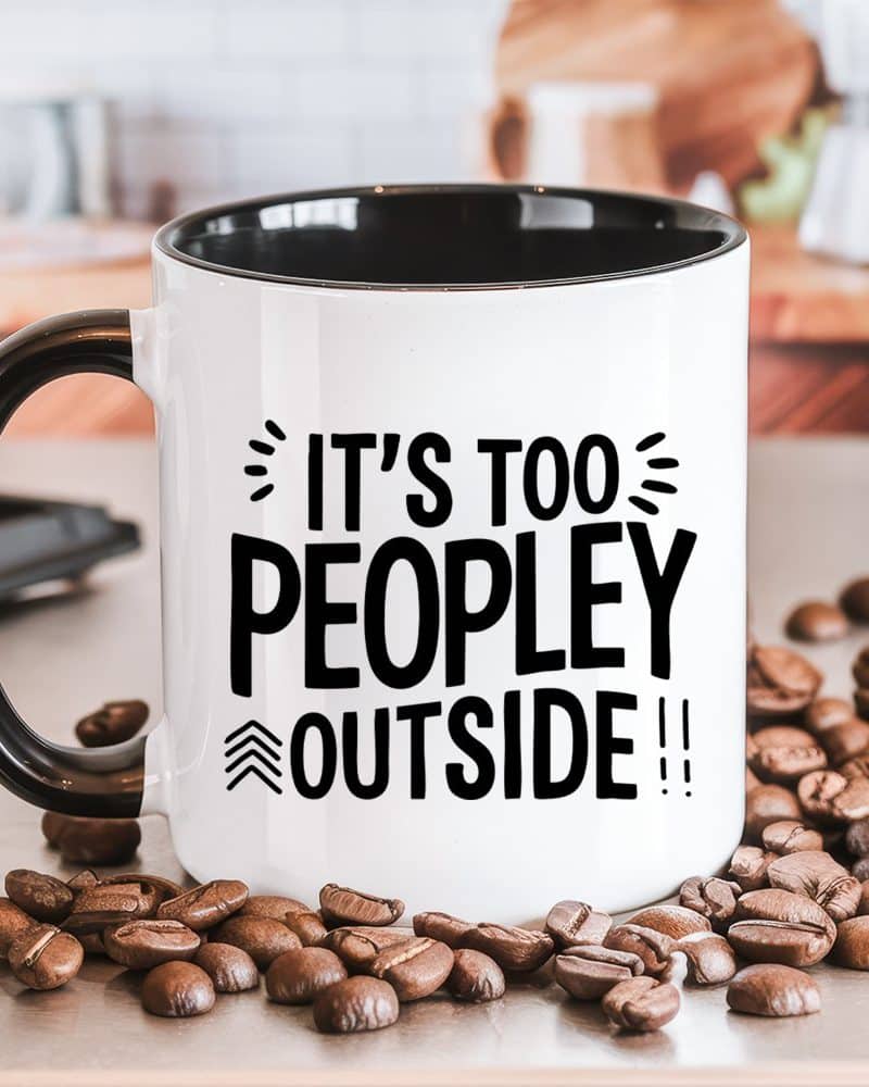 White coffee mug with black interior and handle, displaying the text: Its too people outside. Coffee beans are scattered around the mug.