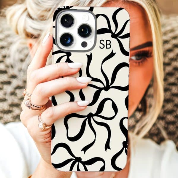 Woman'S Hand Holding An Iphone 12/13 Pro Max Phone Case With A Black Bow Pattern On A Cream Background. The Case Has The Initials Sb.