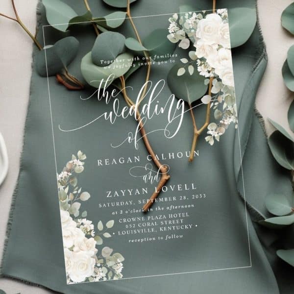 Clear Wedding Invitation With White Flowers And Greenery, Announcing The Wedding Of Reagan Calhoun And Zayyan Lovell.