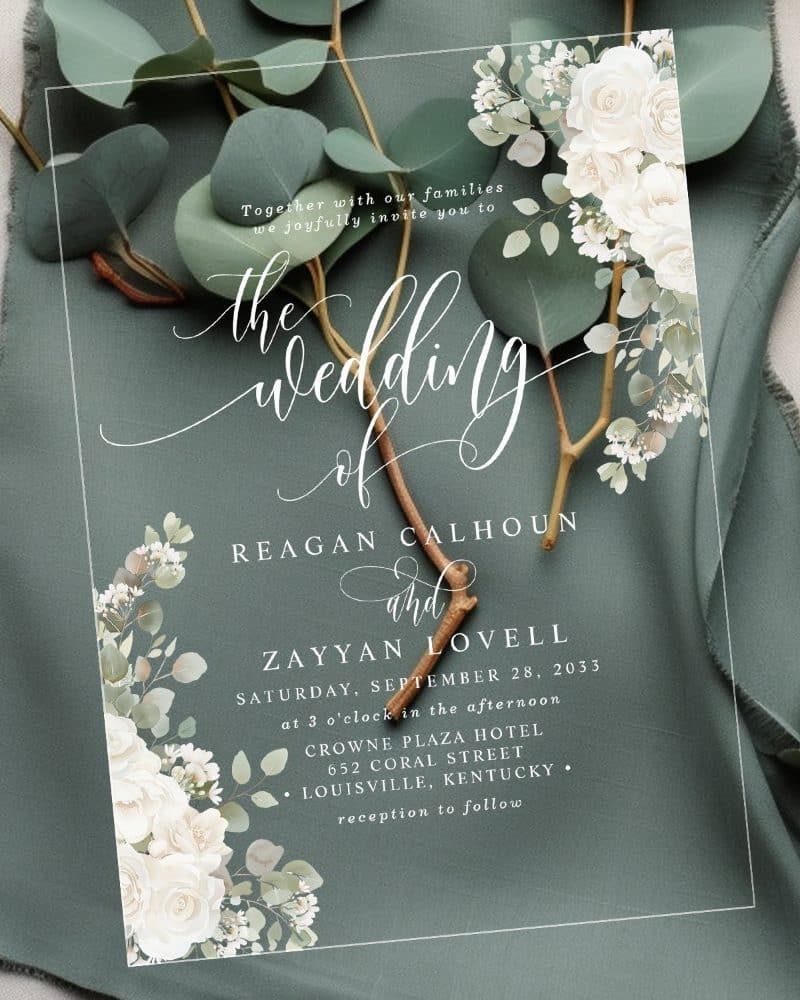 Clear wedding invitation with white flowers and greenery, announcing the wedding of Reagan Calhoun and Zayyan Lovell.