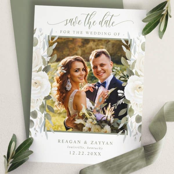 Save The Date Wedding Announcement With A Photo Of The Happy Couple Framed By White Flowers And Green Leaves.