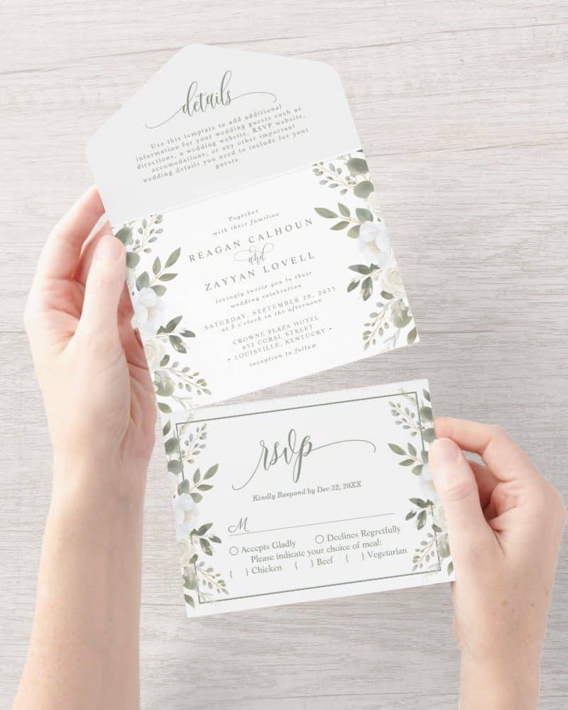Wedding invitation suite with white flowers and green leaves on a white background. The invitation includes the couple's names, wedding date, and location. A separate RSVP card is also shown.
