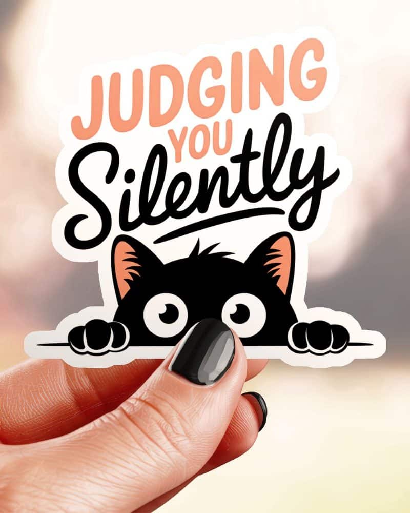 A hand holds a sticker of a black cat peeking over a surface with text that says Judging You Silently.