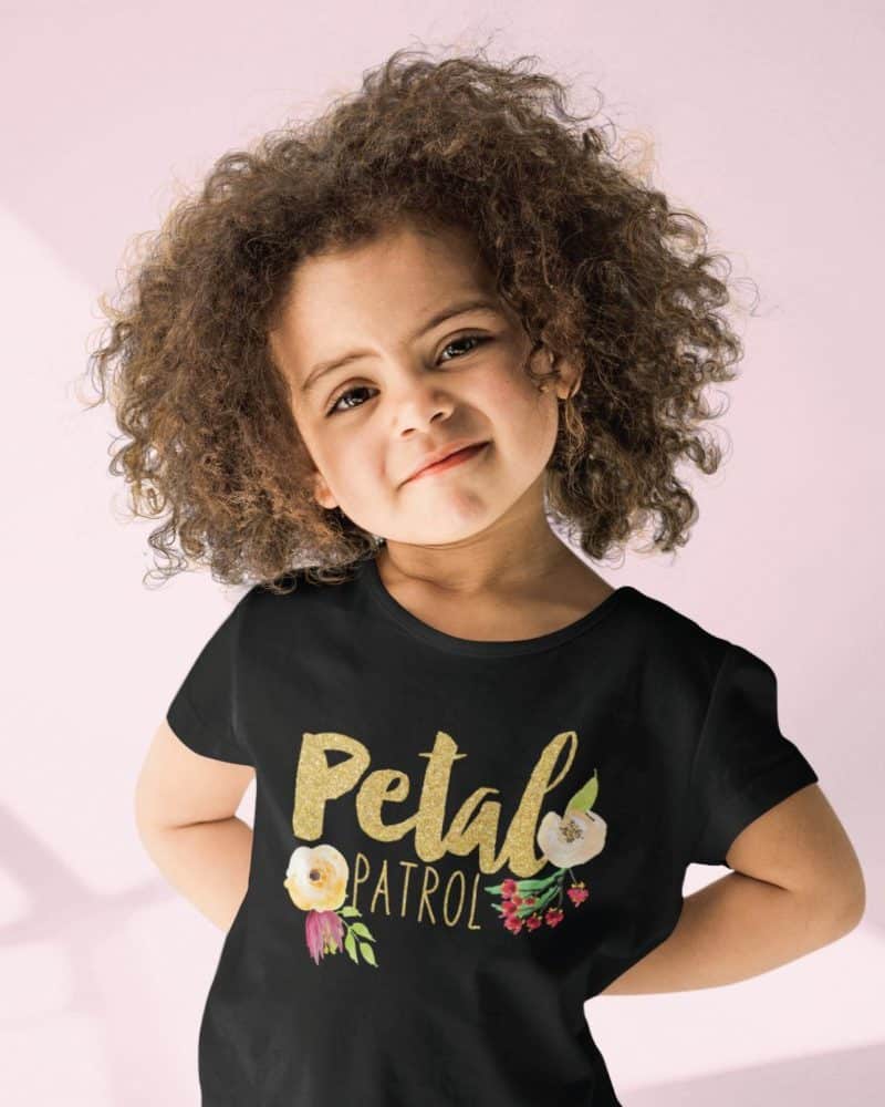 Young girl with curly hair wearing a black t-shirt that says Petal Patrol in gold glitter lettering with floral accents.