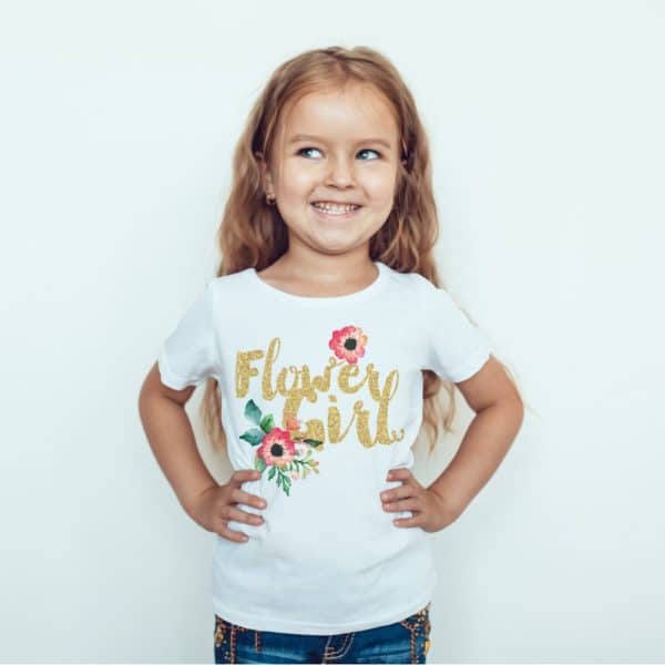 A Young Girl Smiles While Wearing A White T-Shirt That Says Flower Girl In Gold Glitter Lettering With A Small Floral Design.
