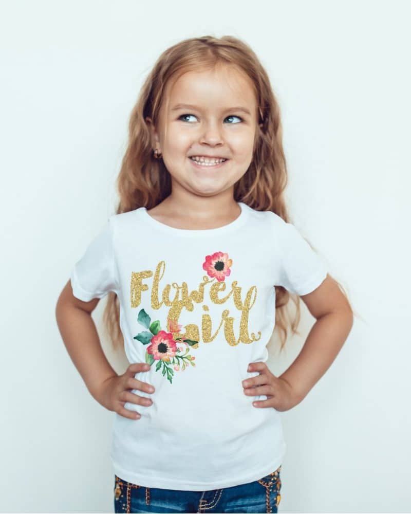A young girl smiles while wearing a white t-shirt that says Flower Girl in gold glitter lettering with a small floral design.