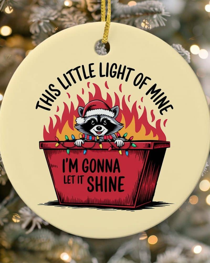Round Christmas ornament featuring a raccoon wearing a Santa hat inside a flaming dumpster decorated with Christmas lights. Text reads, This Little Light of Mine, I'm Gonna Let it Shine.