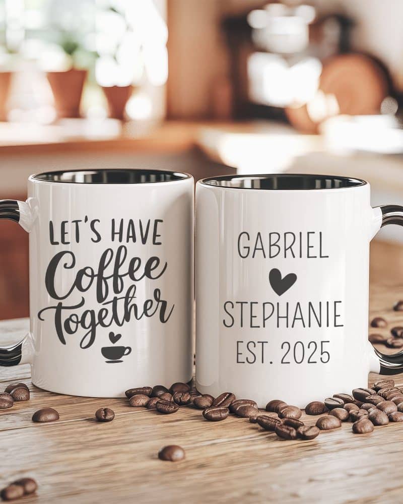 Two white coffee mugs with black handles and rims. One mug says Lets Have Coffee Together, the other says Gabriel Stephanie Est. 2025. Coffee beans are scattered on the table.