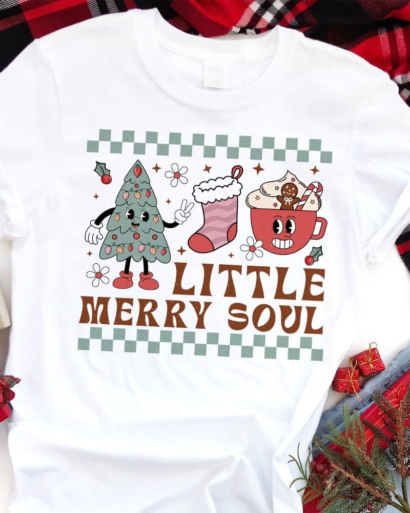 White t-shirt with a retro Christmas design featuring a cartoon Christmas tree, stocking, and mug of hot cocoa. The text Little Merry Soul is below the images.