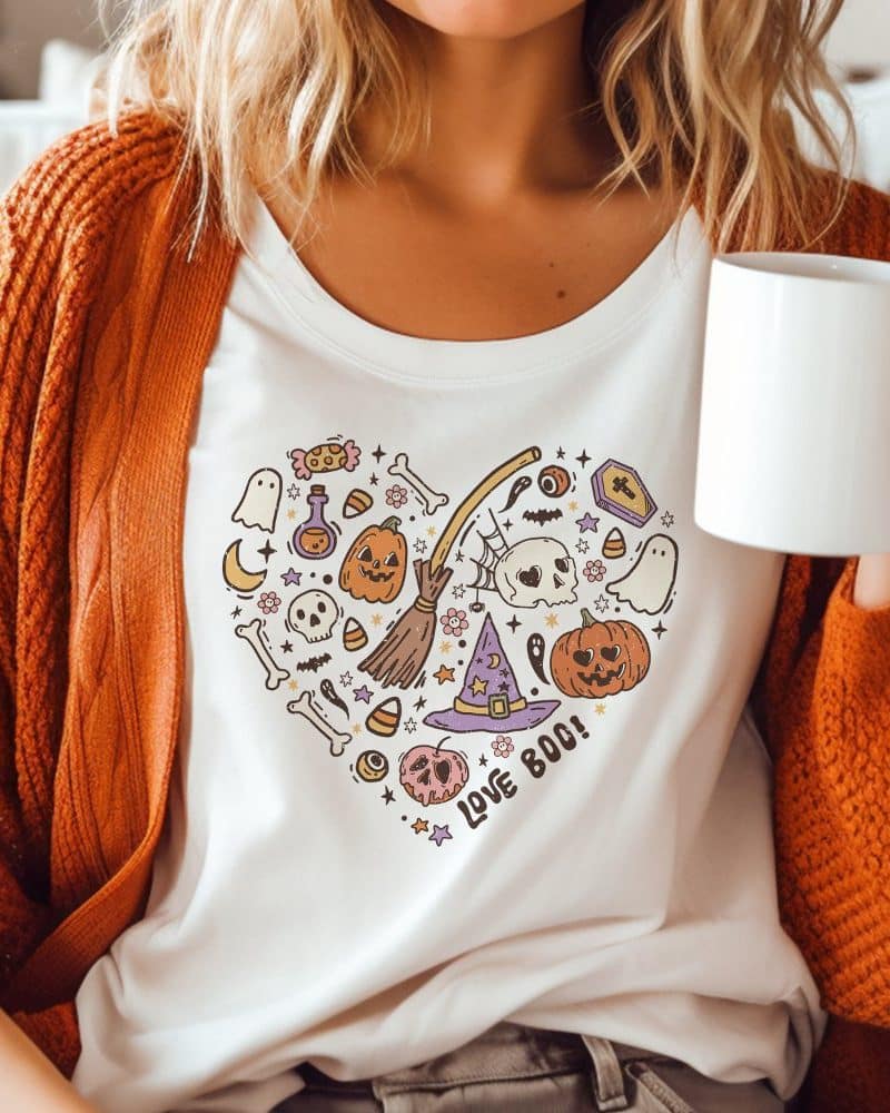 Person wearing a white shirt with a heart-shaped Halloween design featuring pumpkins, ghosts, and a witch's hat.
