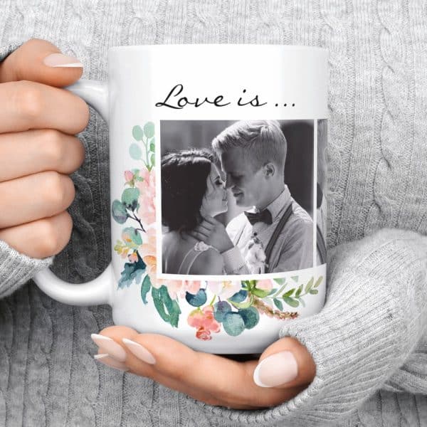 A White Coffee Mug With A Floral Design Surrounding A Black And White Photo Of A Couple Kissing. The Text Love Is... Is Above The Image.