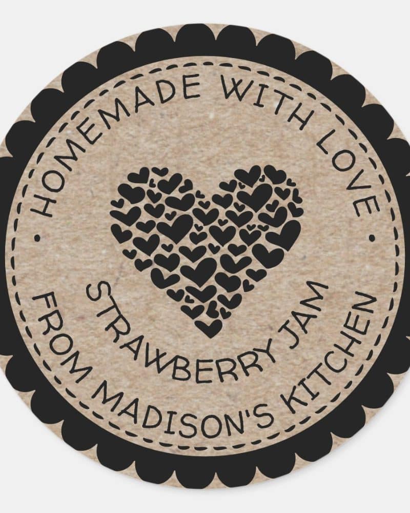 Round label with a black border and scalloped edge, featuring a heart comprised of smaller hearts and text that reads Homemade with Love Strawberry Jam from Madisons Kitchen.