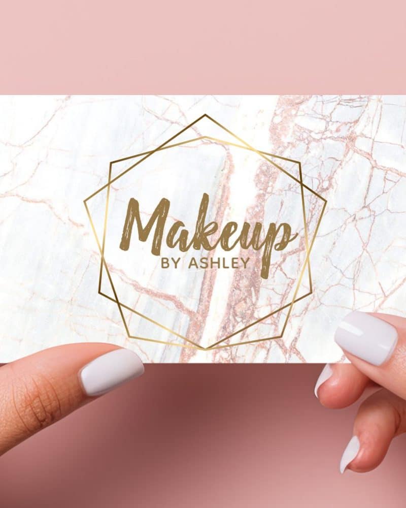 Two hands hold a makeup artist's business card. The card features a marble background with a gold geometric frame and the text Makeup By Ashley.