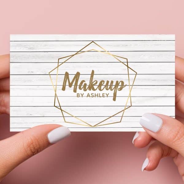 Two Hands Hold A Business Card With A White Wood Grain Background. A Gold Geometric Frame Encloses The Text Makeup By Ashley.